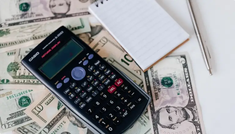 A calculator, notepad, and pen on top of dollar bills.